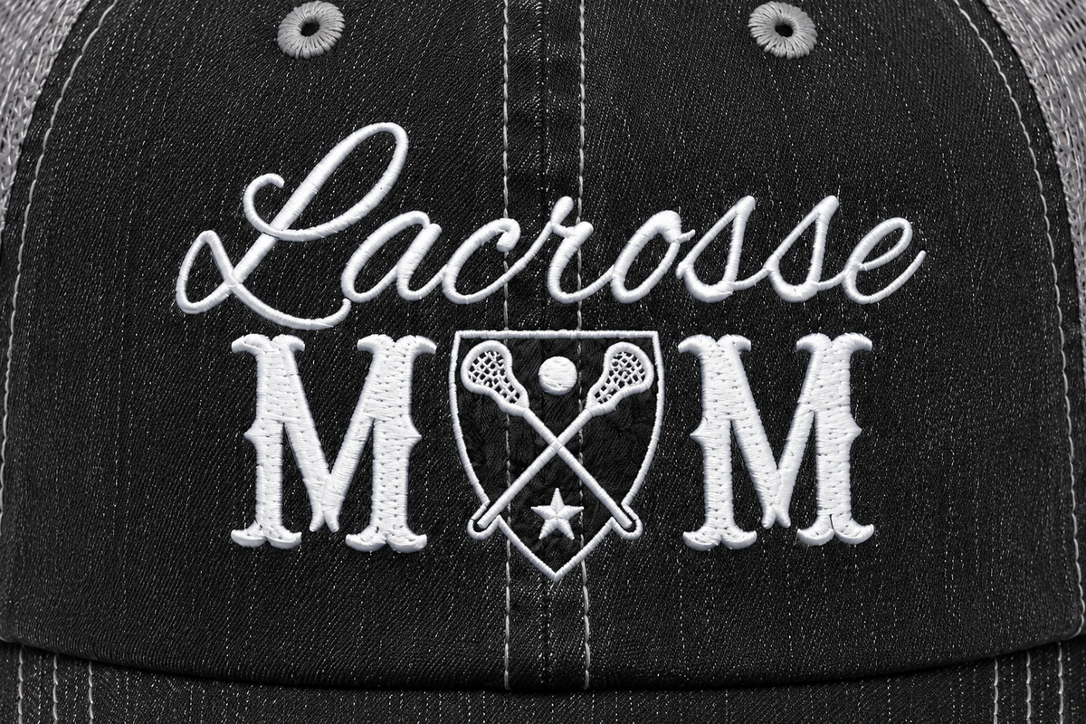 Close-up of embroidered "Lacrosse MOM" crest design on a washed black distressed trucker hat front panel.
