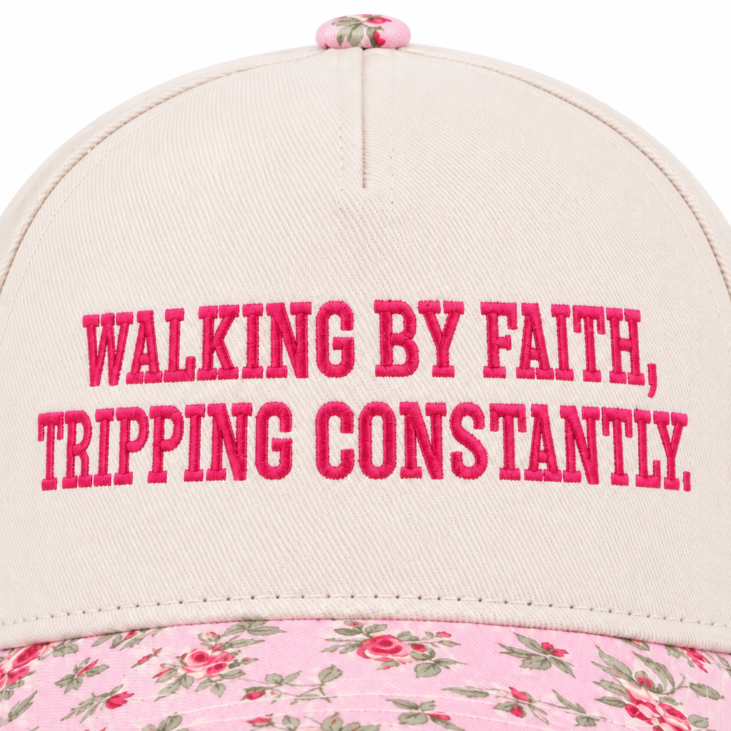 Close-up of Walking By Faith Tripping Constantly Baseball Cap in Floral showing hot pink raised block embroidered lettering reading Walking By Faith Tripping Constantly on cream structured cotton crown with pink floral bill edge visible at bottom on white background
