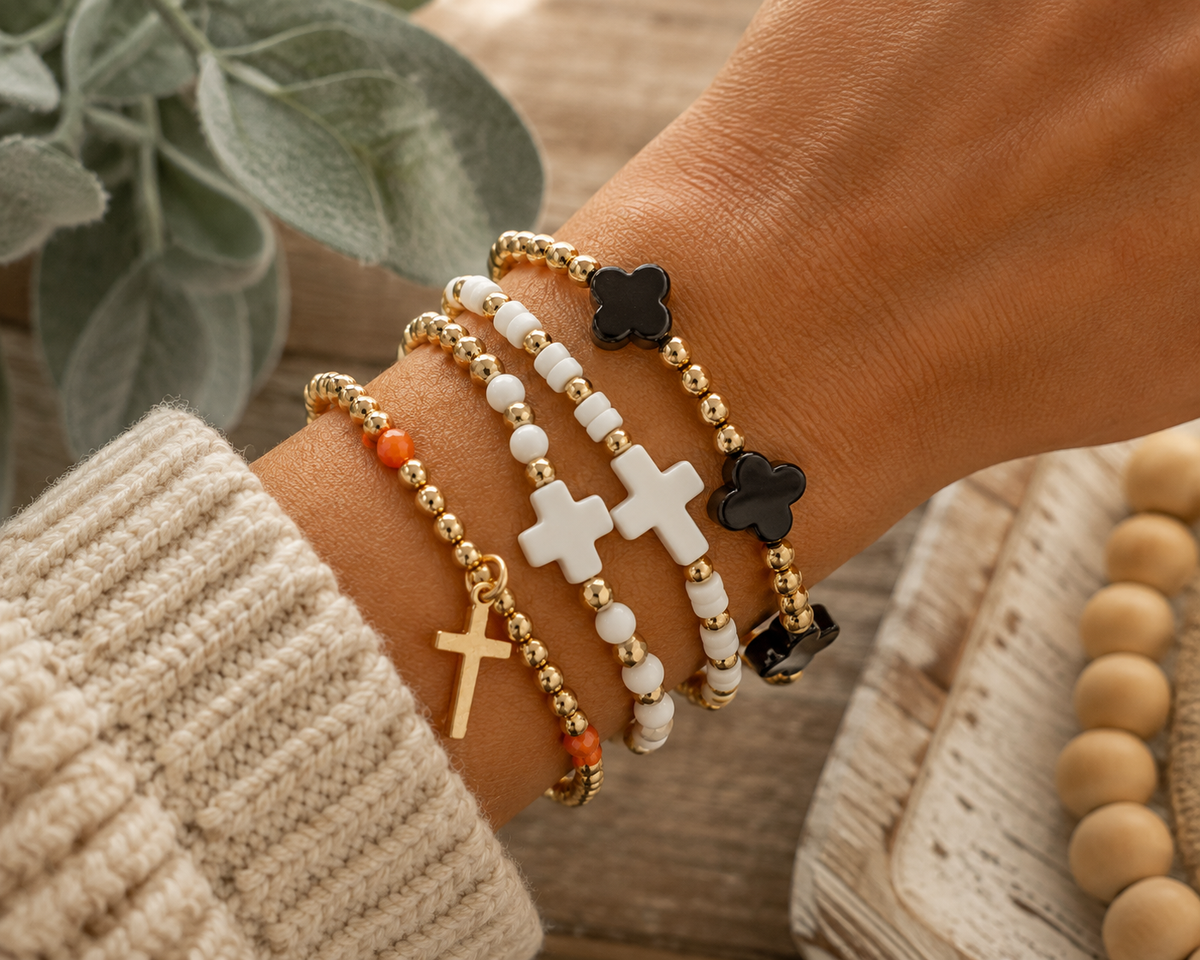 Gold and orange cross bracelet stacked with white cross bead bracelet, white seed bead bracelet, and black clover gold bracelet on wrist with cream knit sweater
