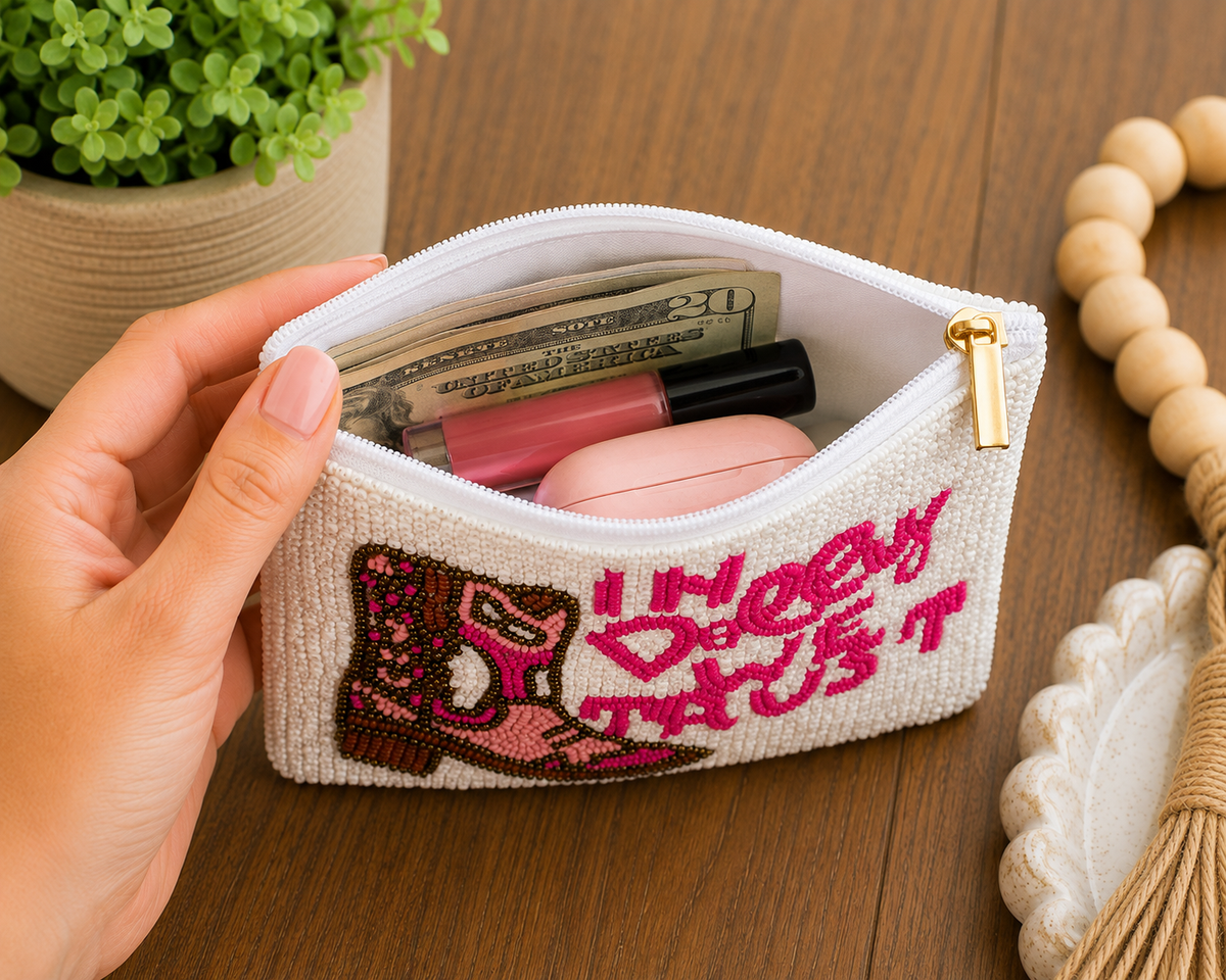 A hand holds an open white beaded zip pouch with a gold zipper, showing it filled with cash, lip gloss, and a pink case, set on a wooden tabletop with a potted plant in the background.
