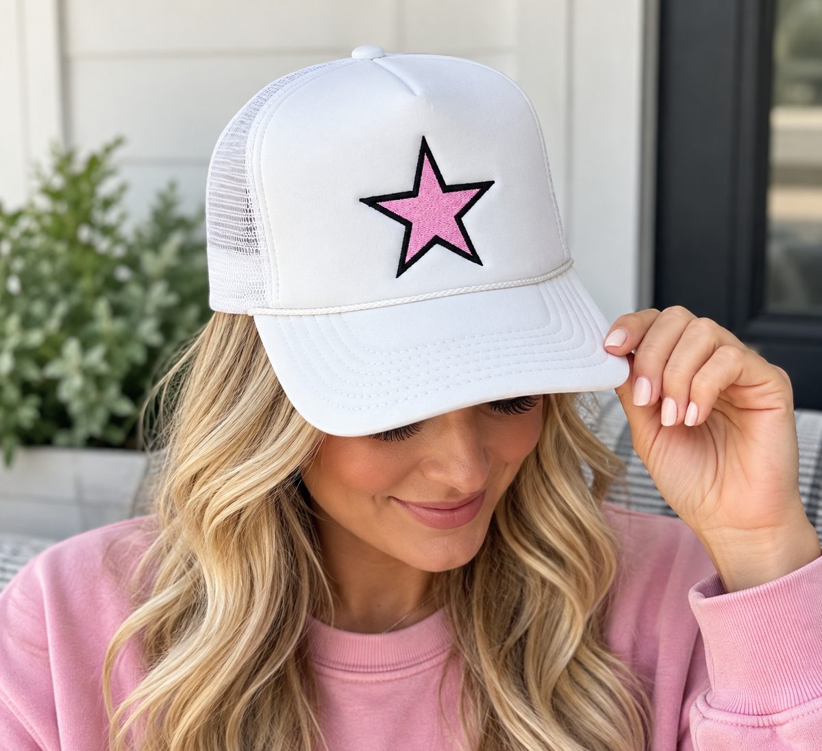 Woman wearing Pink Star Trucker Hat with white front and white mesh back showing pink embroidered star with black outline with pink sweatshirt and outdoor porch backdrop
