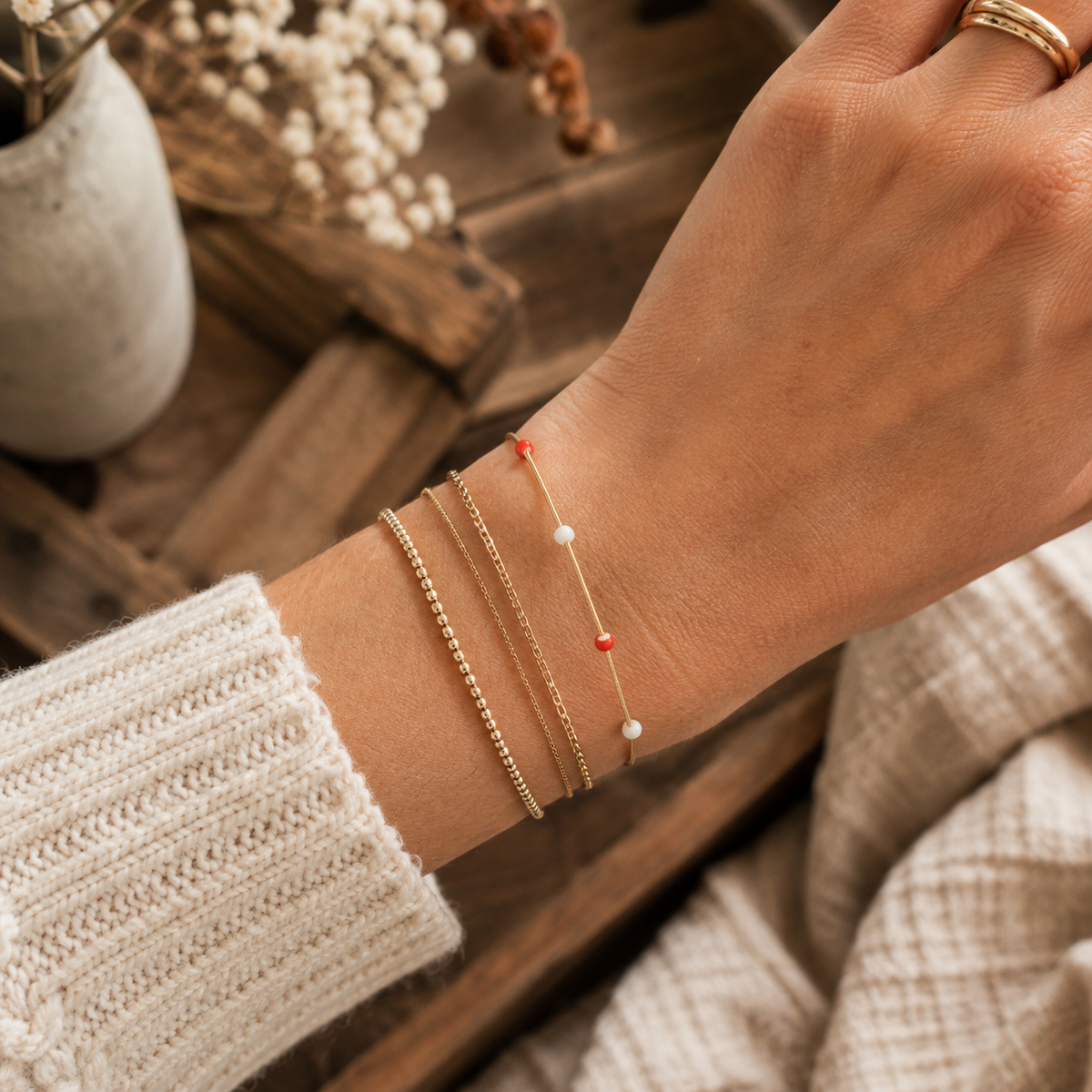 Gold coil bracelet with red and white beads shown stacked with gold chain bracelets on wrist with cream knit sleeve, rustic wood crate, and dried floral backdrop
