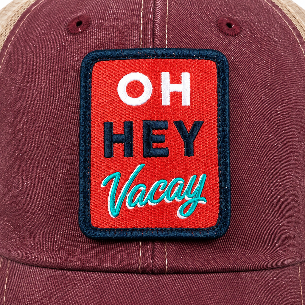Close-up of the Oh Hey Vacay embroidered patch on a burgundy trucker hat, featuring bold white OH, navy HEY, and teal script Vacay text on a red background with a navy border.