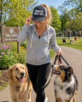A woman walking two dogs on a paved path in a park, wearing a gray zip-up hoodie, black leggings, and a black trucker hat that reads "Pawsitive Vibes Only."
