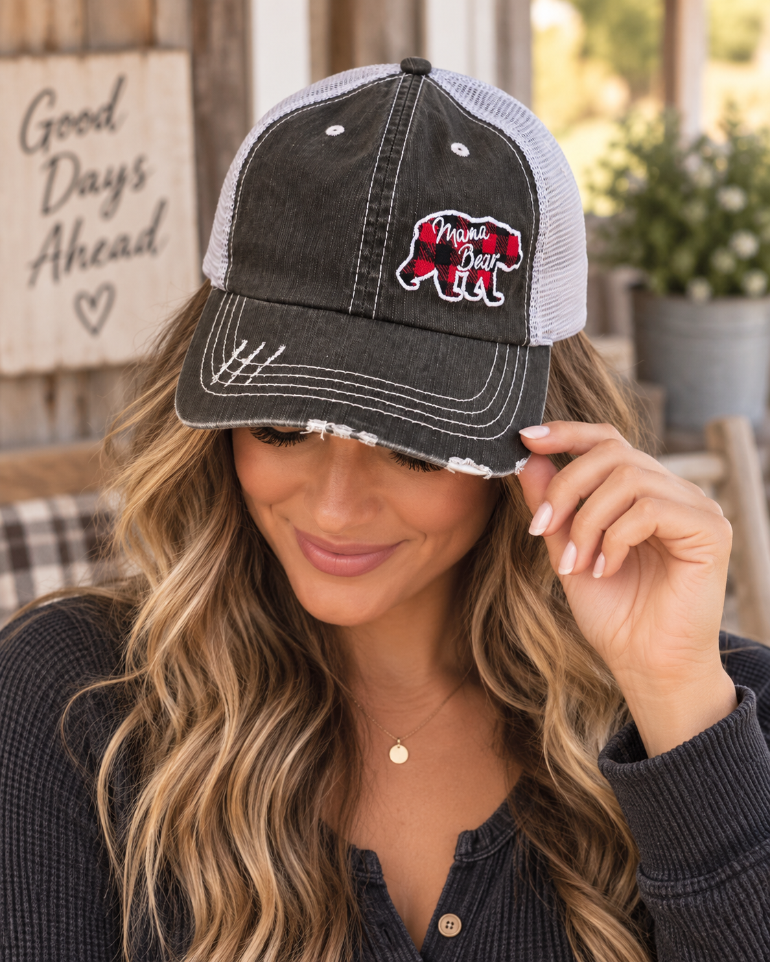 Woman wearing Mama Bear Trucker Hat with distressed black front and white mesh back showing red and black buffalo plaid bear patch with white Mama Bear script with dark navy henley and rustic backdrop
