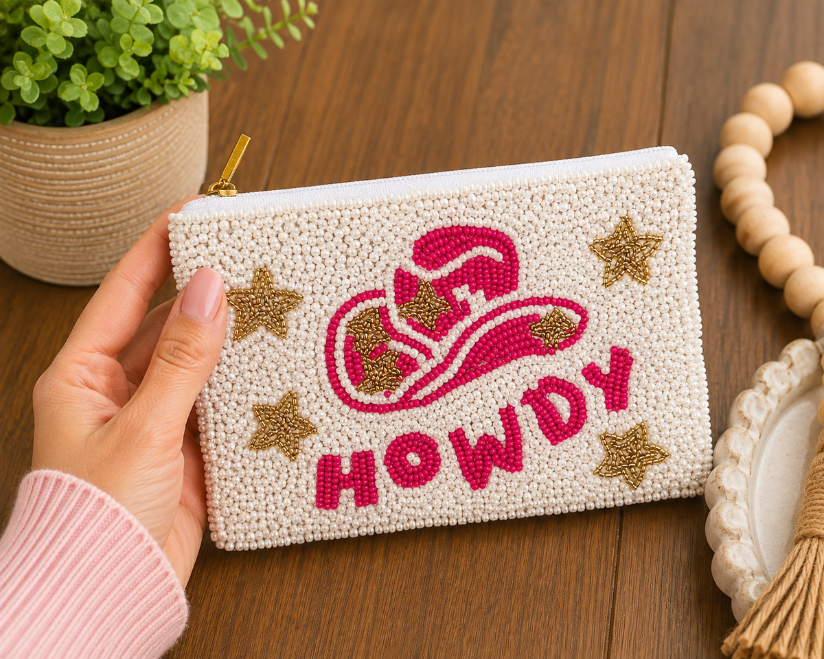 A hand holds a small zippered pouch decorated with a pink cowboy hat and the word "HOWDY" in pink beads, surrounded by gold star accents on a cream beaded background. The pouch is photographed on a wooden table with decorative props including a potted plant, wooden beads, and a tassel.