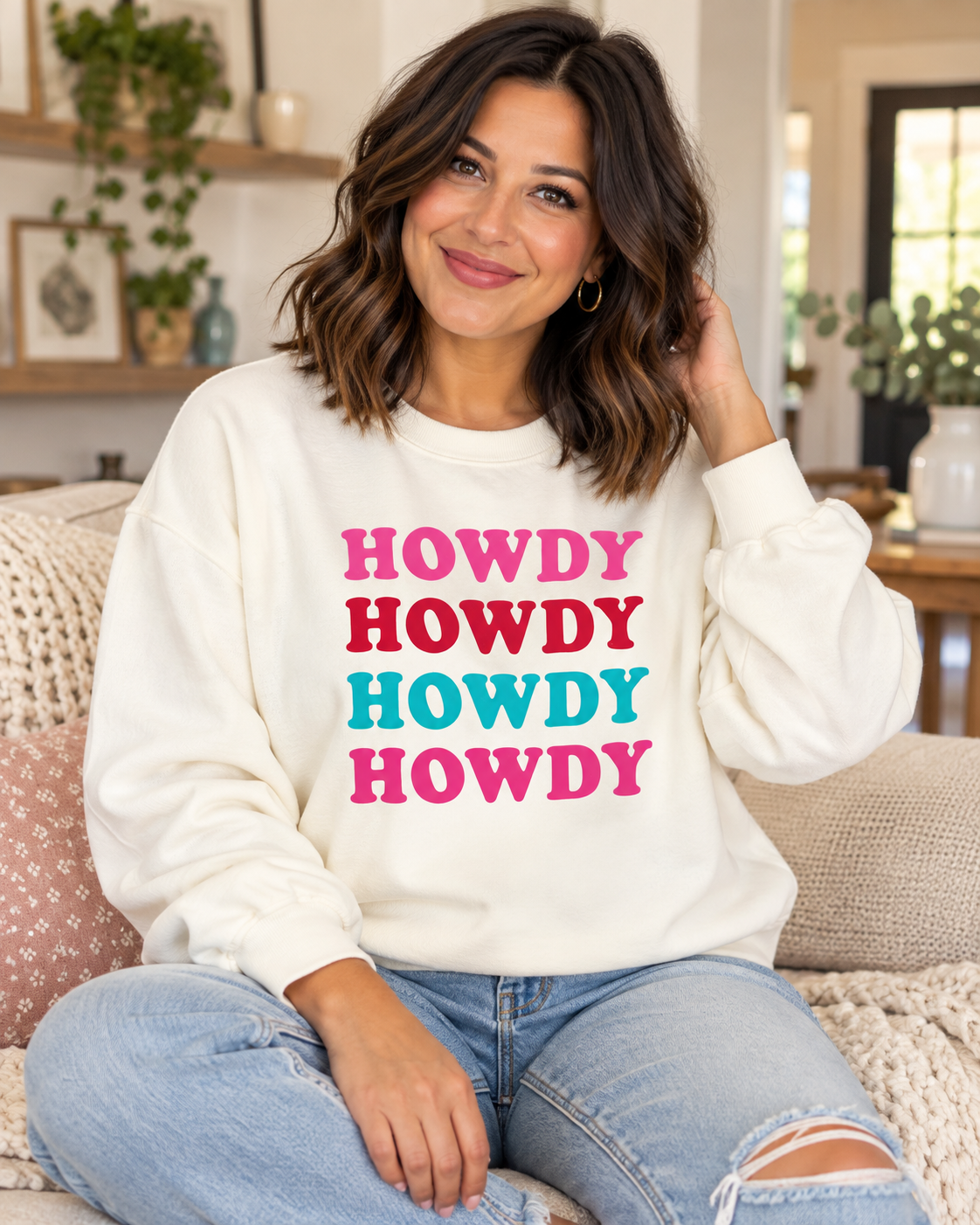 Seated model wearing a cream HOWDY crewneck sweatshirt with colorful stacked lettering and blue jeans
