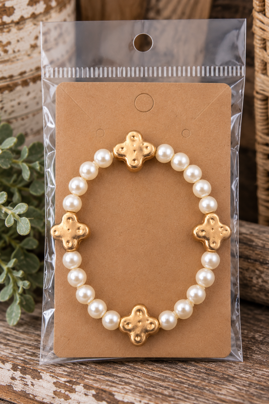 Pearl beaded bracelet with gold clover accents on kraft card in clear packaging on rustic wood surface with chippy white round box and greenery backdrop

