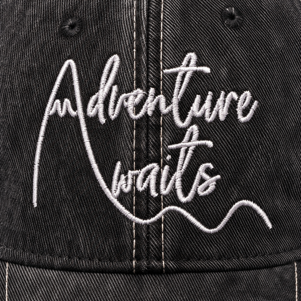 Close-up of white Adventure Awaits script embroidery on distressed black trucker hat
