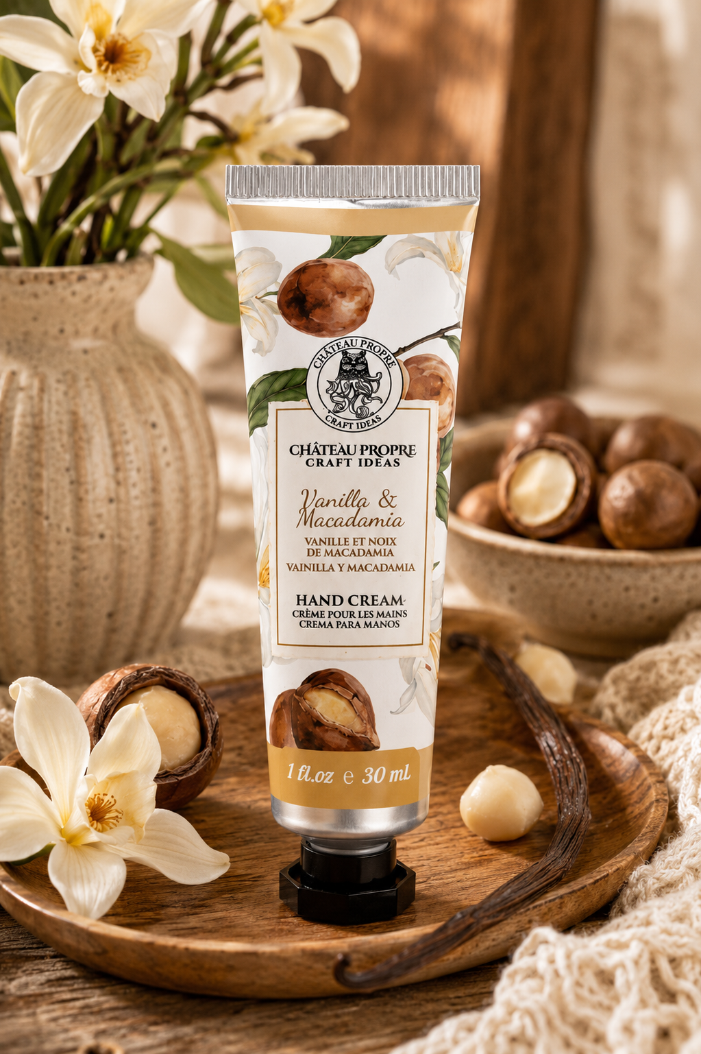 A squeeze tube of hand cream from Chateau Propre Craft Ideas, scented Vanilla & Macadamia, standing on a wooden tray surrounded by macadamia nuts, white flowers, and a textured blanket.