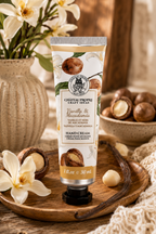 A squeeze tube of hand cream from Chateau Propre Craft Ideas, scented Vanilla & Macadamia, standing on a wooden tray surrounded by macadamia nuts, white flowers, and a textured blanket.