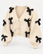Angled view of the tan knit cardigan showing black bows across the front and sleeves, dropped shoulders, ribbed cuffs, and cropped hem
