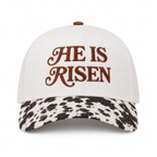 He Is Risen Baseball Cap in Cow front view showing cream cotton crown with brown decorative script He Is Risen embroidery, black and white cow print bill, and brown top button on white background
