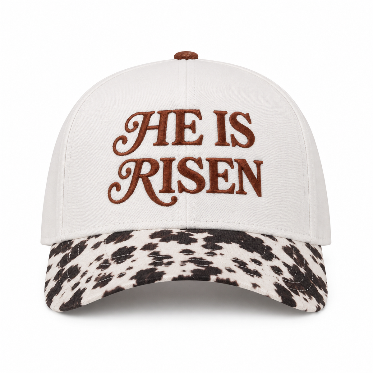 He Is Risen Baseball Cap in Cow front view showing cream cotton crown with brown decorative script He Is Risen embroidery, black and white cow print bill, and brown top button on white background
