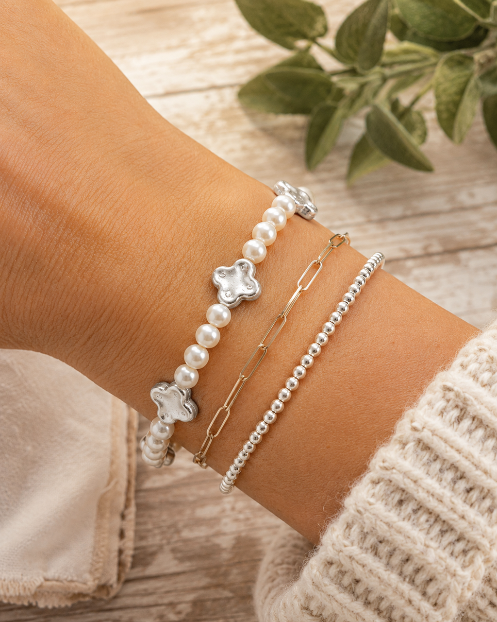 Pearl and silver clover bracelet stacked with gold paperclip chain bracelet and silver ball bead bracelet on wrist with cream knit sweater and whitewashed wood backdrop
