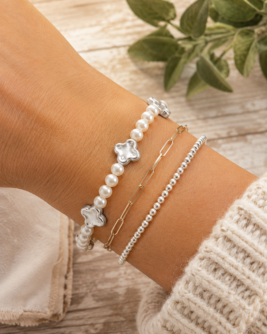 Pearl and silver clover bracelet stacked with gold paperclip chain bracelet and silver ball bead bracelet on wrist with cream knit sweater and whitewashed wood backdrop
