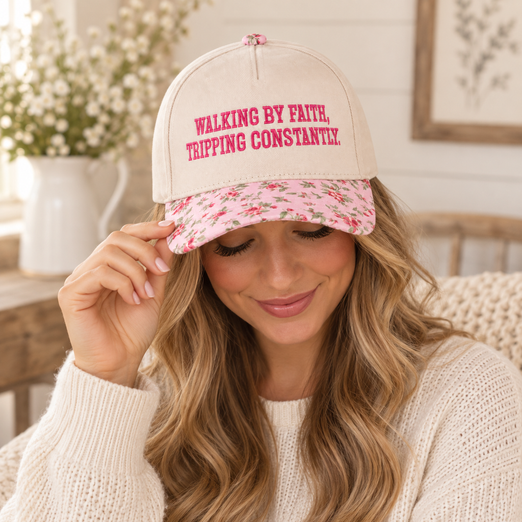 Blonde woman with wavy hair in cream chunky knit sweater smiling down while tilting brim of Walking By Faith Tripping Constantly Baseball Cap in Floral showing cream crown with hot pink embroidered lettering and pink floral bill, seated indoors with white shiplap wall and white vase with white florals in background
