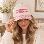 Blonde woman with wavy hair in cream chunky knit sweater smiling down while tilting brim of Walking By Faith Tripping Constantly Baseball Cap in Floral showing cream crown with hot pink embroidered lettering and pink floral bill, seated indoors with white shiplap wall and white vase with white florals in background
