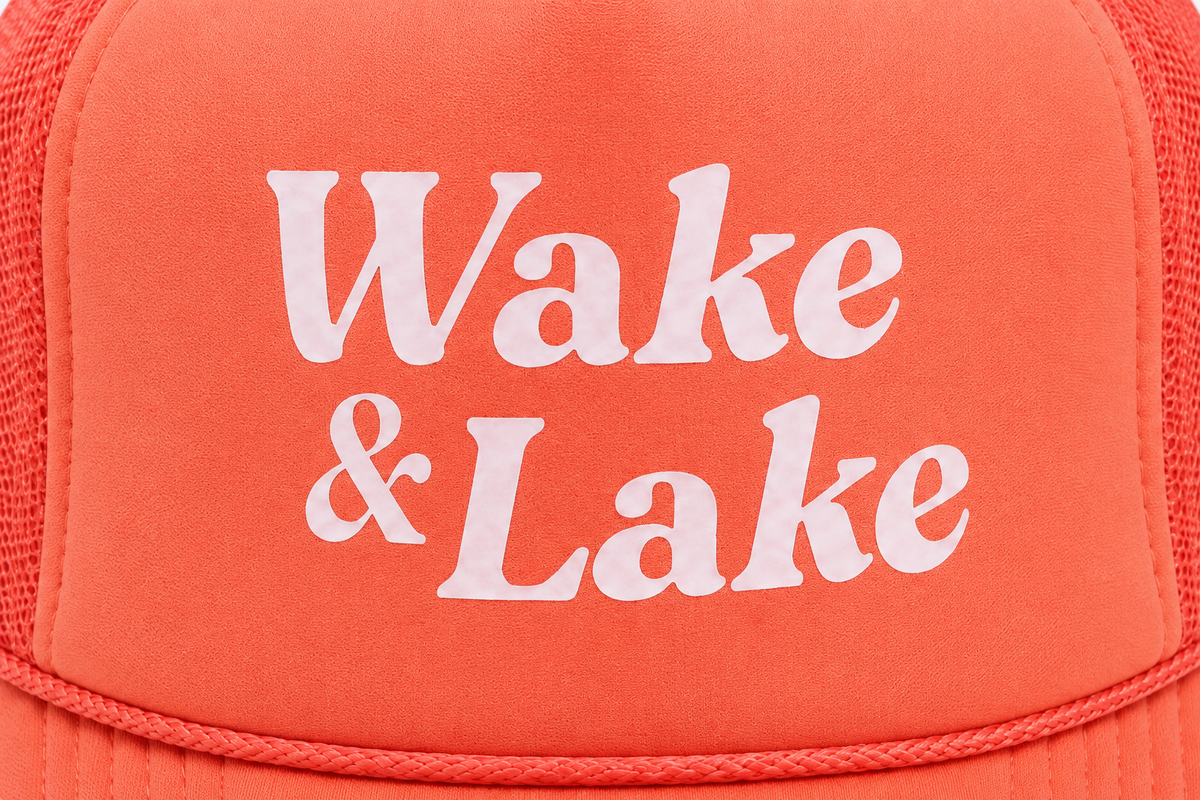 Close-up of white retro "Wake & Lake" printed lettering on a coral-orange foam trucker hat front panel.
