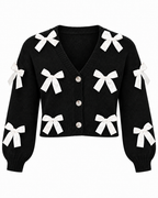 Black Satin Bow Knit Cardigan front view showing black knit body with white satin ribbon bows appliquéd on front and sleeves, deep V-neckline, pearl dome button placket, and elasticized ribbed cuffs on white background
