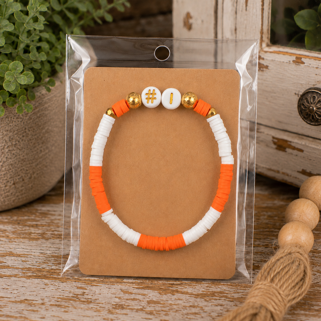 Orange and white #1 beaded stretch bracelet on kraft card in clear packaging displayed on rustic wood surface with wood bead garland
