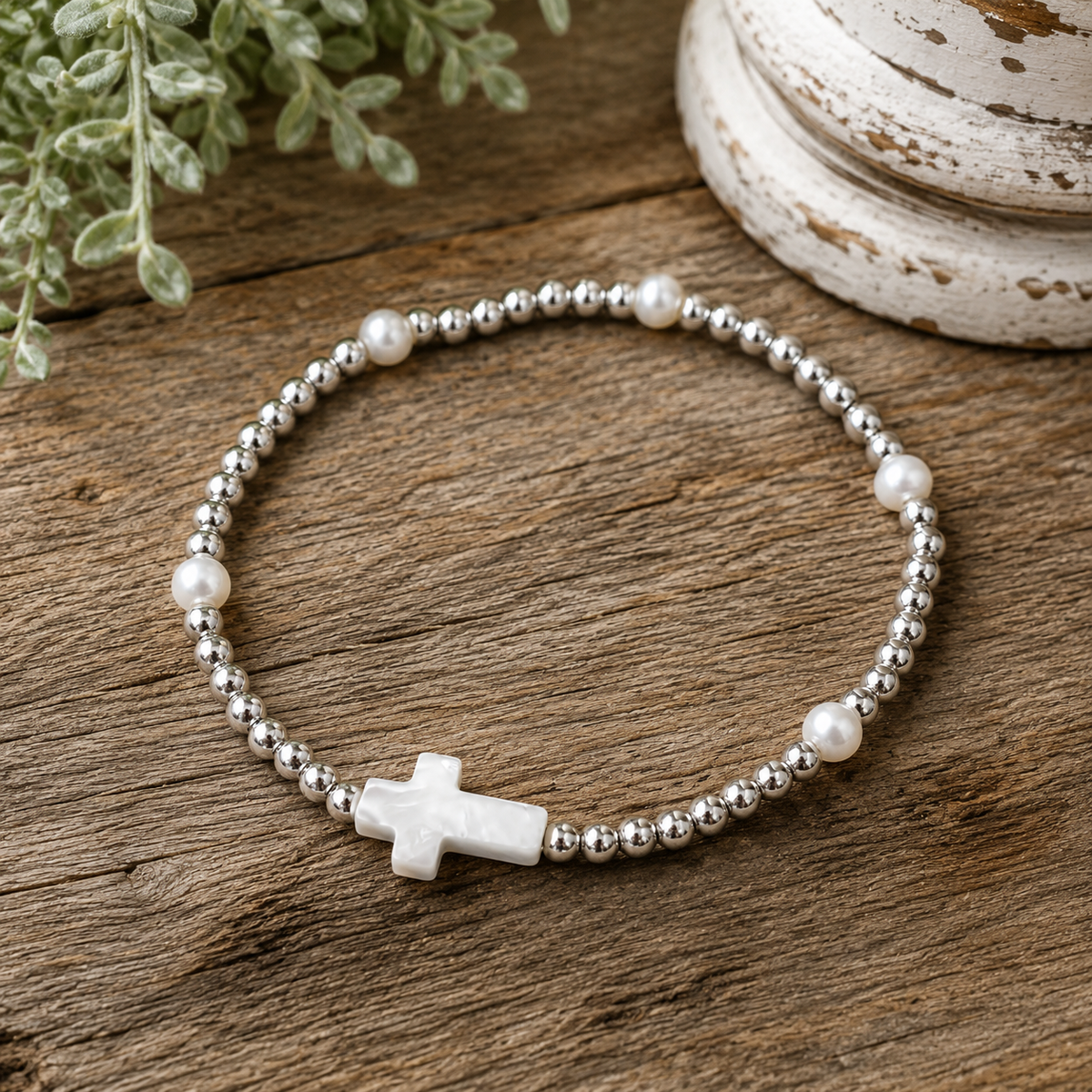 Silver beaded cross bracelet with pearl accents laid flat in a circle on rustic wood surface with chippy white pedestal and greenery backdrop
