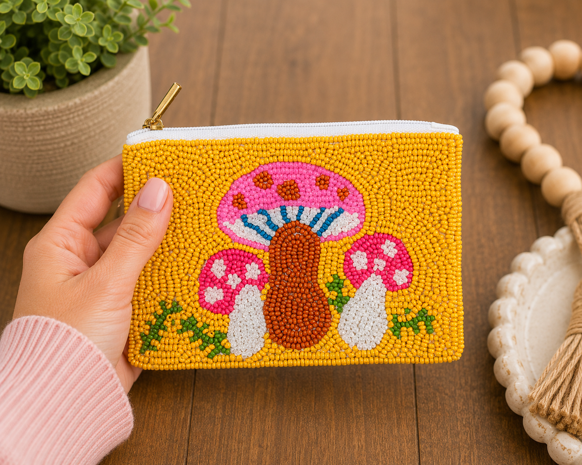 A hand holds a yellow beaded coin purse with a colorful mushroom design, set against a wooden table background with a potted plant and a macrame decor piece.