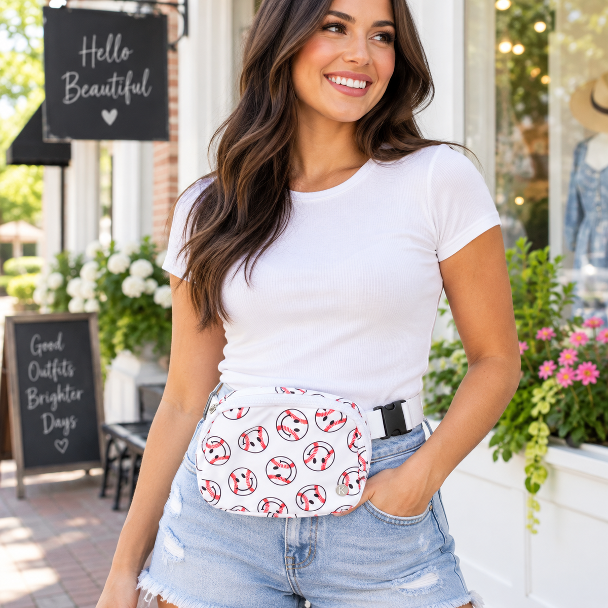 Smiling woman in white tee and denim shorts wearing Baseball Smiley Belt Bag at the waist showing white nylon with allover red and black baseball smiley print and adjustable white strap with black buckle in front of Hello Beautiful boutique chalkboard and Good Outfits Brighter Days sign with pink and white flowers
