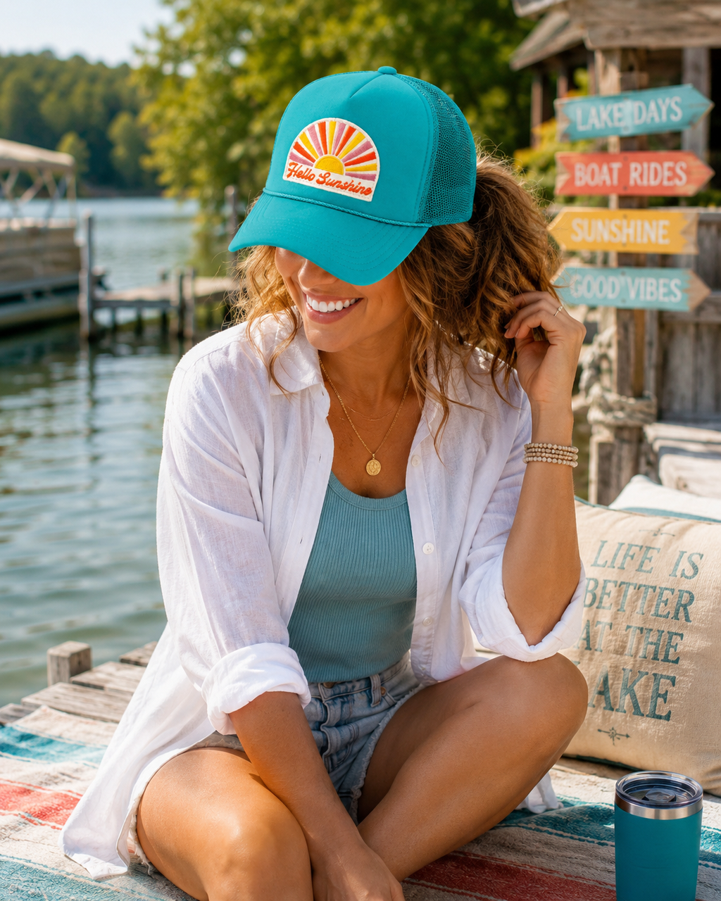 A woman sitting on a wooden dock by the water wearing a teal Hello Sunshine trucker hat with a retro sunburst patch, white button-up, and denim shorts.
