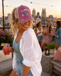 A woman on a rooftop patio at sunset wearing a dusty rose and beige trucker hat with 'I'll Bring the Bad Decisions' embroidered text, smiling and holding a drink with a city skyline in the background.