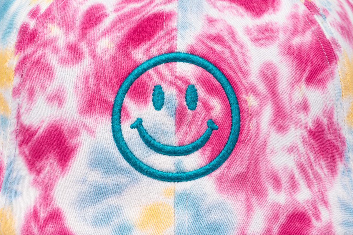 Close-up of embroidered teal smiley face on a vibrant pink, blue, and yellow tie-dye cap front panel.

