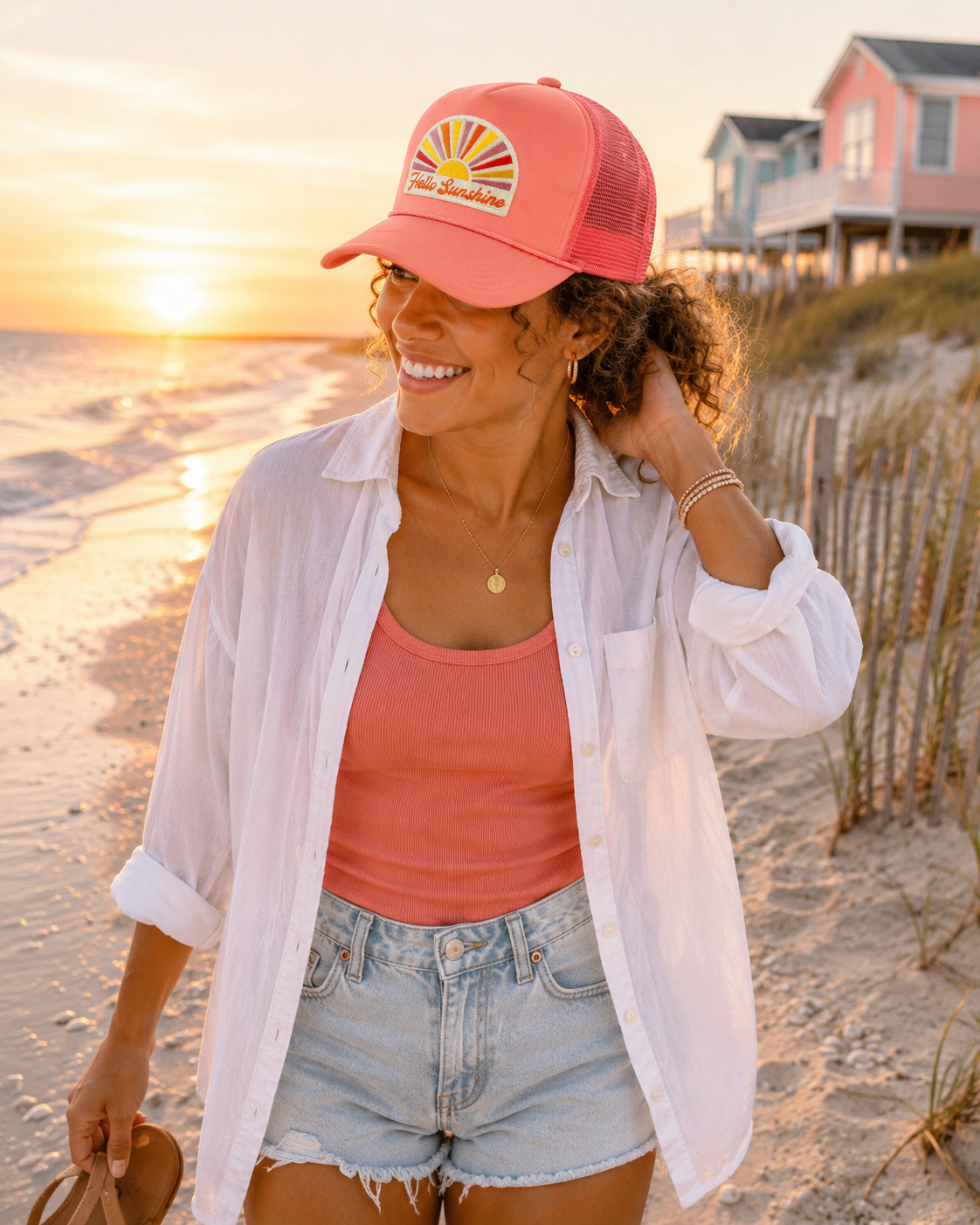 A woman on a sandy beach at sunset wearing a coral pink trucker hat with a retro sunburst "Hello Sunshine" embroidered patch, paired with a white button-down and denim shorts.
