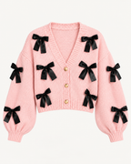 Front view of the pink cropped cardigan showing black ribbon bows, a ribbed V-neckline, three gold-tone buttons, and long sleeves
