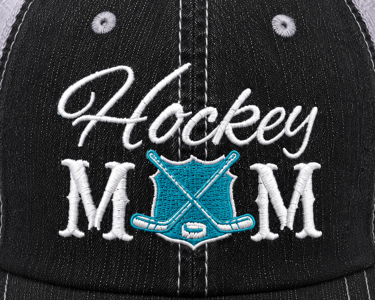 Close-up of white embroidered "Hockey MOM" lettering with crossed hockey sticks and puck detail on a distressed black trucker hat front panel.
