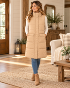 Woman wearing a tan long quilted puffer vest over a cream long-sleeve top with jeans and tan ankle boots, showing the stand collar and front pockets
