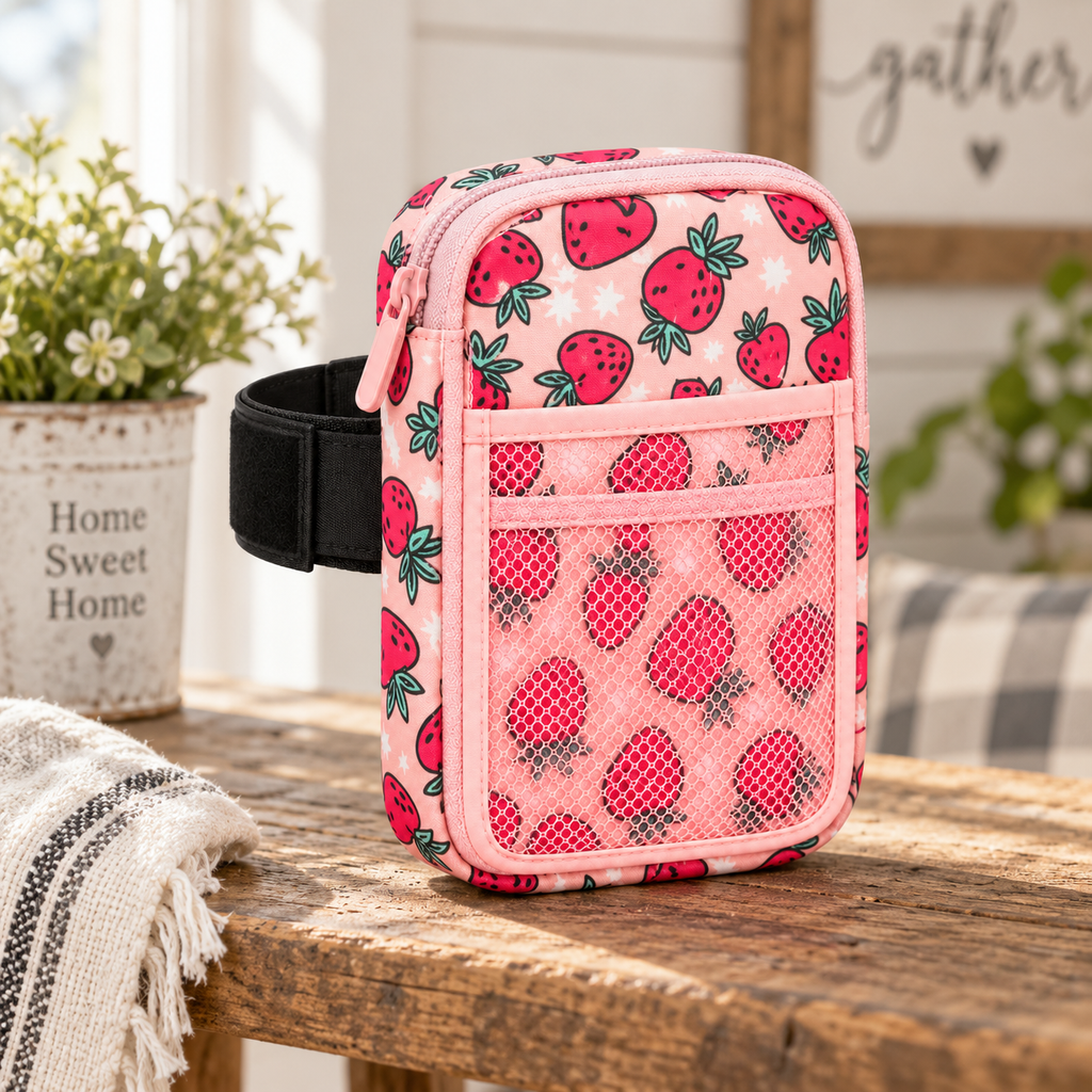 Strawberry Tumbler Pouch on rustic wood farmhouse table with black elastic tumbler strap showing light pink all-over strawberry and star print and front mesh pocket beside white pot of flowers, striped throw, and Home Sweet Home and gather signs
