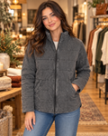 Brunette woman smiling in charcoal garment washed quilted jacket unzipped over white tee with blue jeans, in warm boutique interior with clothing racks and fall decor

