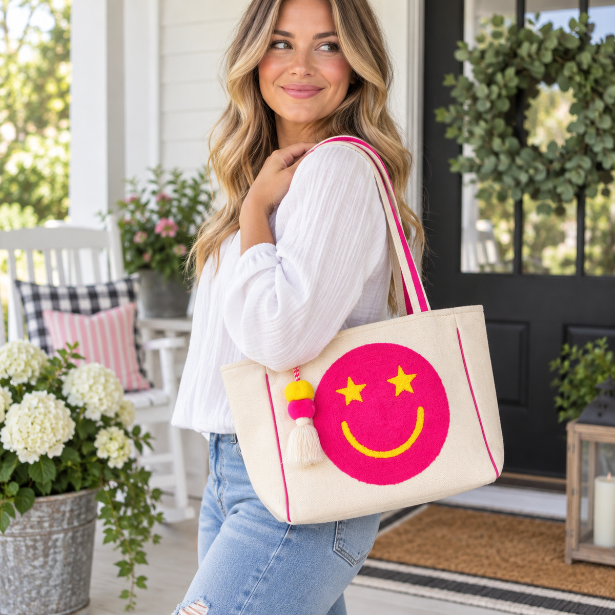 Woman in white blouse and jeans carrying Pink Star Smiley Canvas Tote Bag on shoulder showing natural canvas with oversized hot pink chenille smiley face, pink-striped handles, and pom-pom tassel charm on white farmhouse porch with black front door, round topiaries, and white hydrangeas
