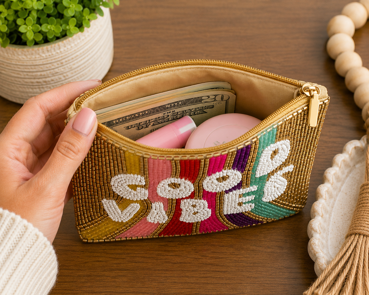 A hand holds an open zippered cosmetic pouch on a wooden tabletop. The pouch has a gold-tone zipper, a beige interior, and colorful embroidered stripes in pink, purple, green, and blue.