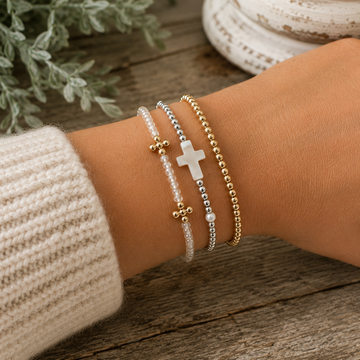 Clear gold cross bracelet stacked with silver and pearl cross bracelet and gold beaded bracelet on wrist with cream knit sweater and rustic wood backdrop
