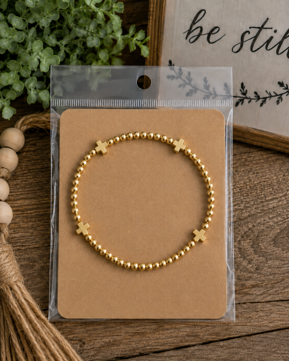 Gold cross beaded stretch bracelet on kraft card in clear packaging on rustic wood surface with wood bead garland, jute tassel, greenery, and be still sign
