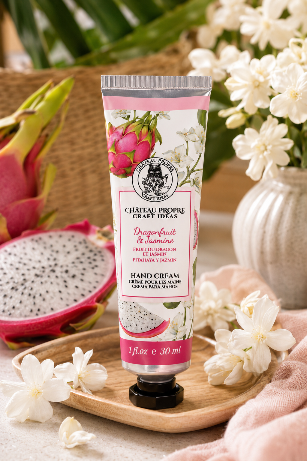 A squeeze tube of Château Propre Craft Ideas Dragonfruit & Jasmine Hand Cream stands on a wooden tray, surrounded by dragonfruit slices, white flowers, and soft pink fabric in a styled setting.