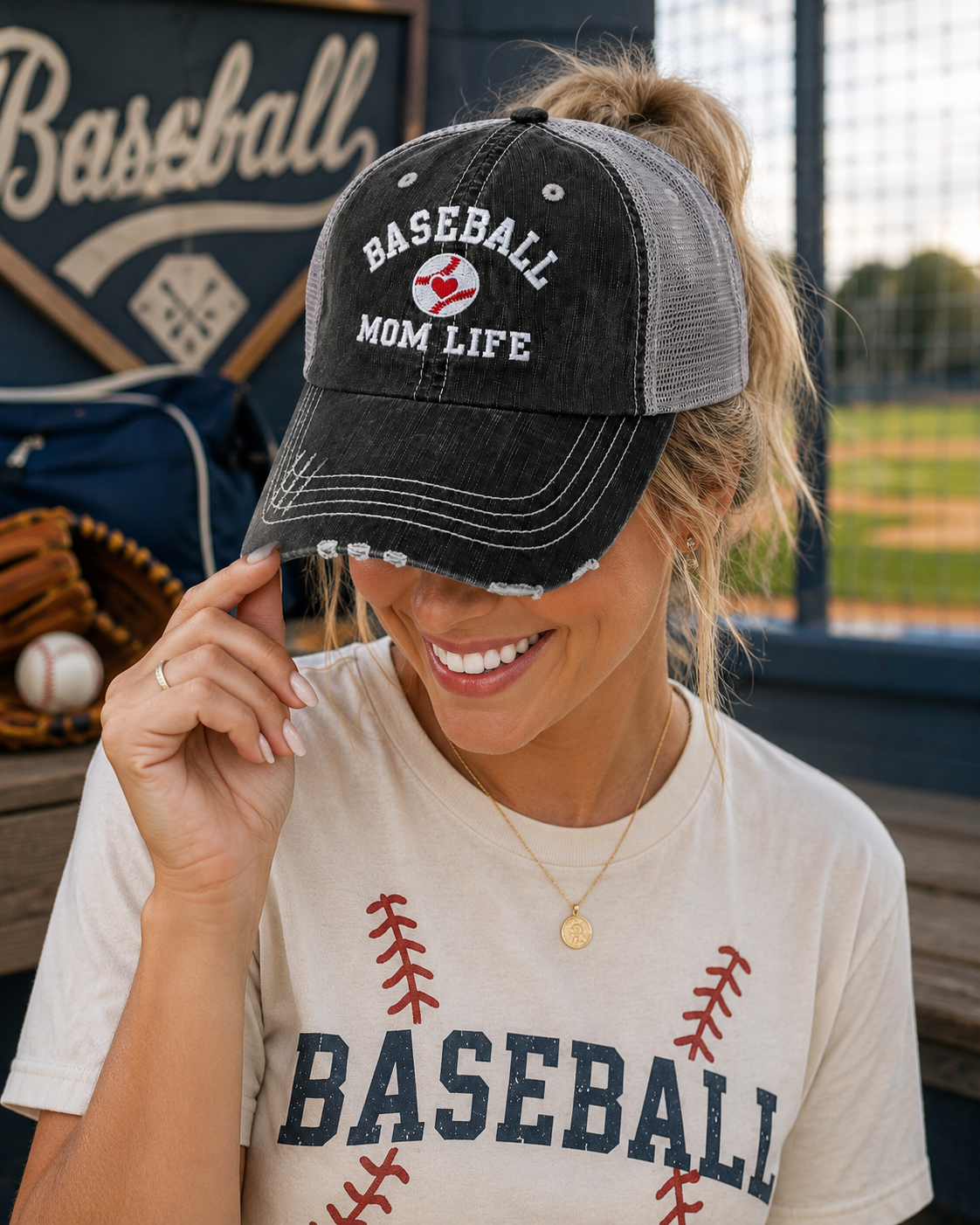 A woman at a baseball field wearing a distressed black and gray trucker hat that says 'BASEBALL MOM LIFE' with a small red heart logo, paired with a cream baseball graphic t-shirt.