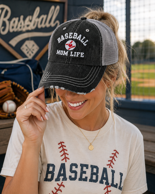 A woman at a baseball field wearing a distressed black and gray trucker hat that says 'BASEBALL MOM LIFE' with a small red heart logo, paired with a cream baseball graphic t-shirt.