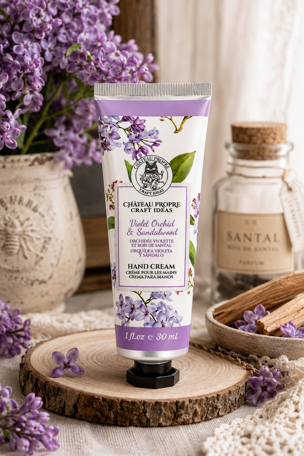 A tube of Château Propre Craft Ideas Violet Orchid & Sandalwood Hand Cream is displayed on a rustic wood slice. The scene is styled with purple flowers, a small glass bottle of Santal, and a bowl of wood chips, creating a warm, natural aesthetic.