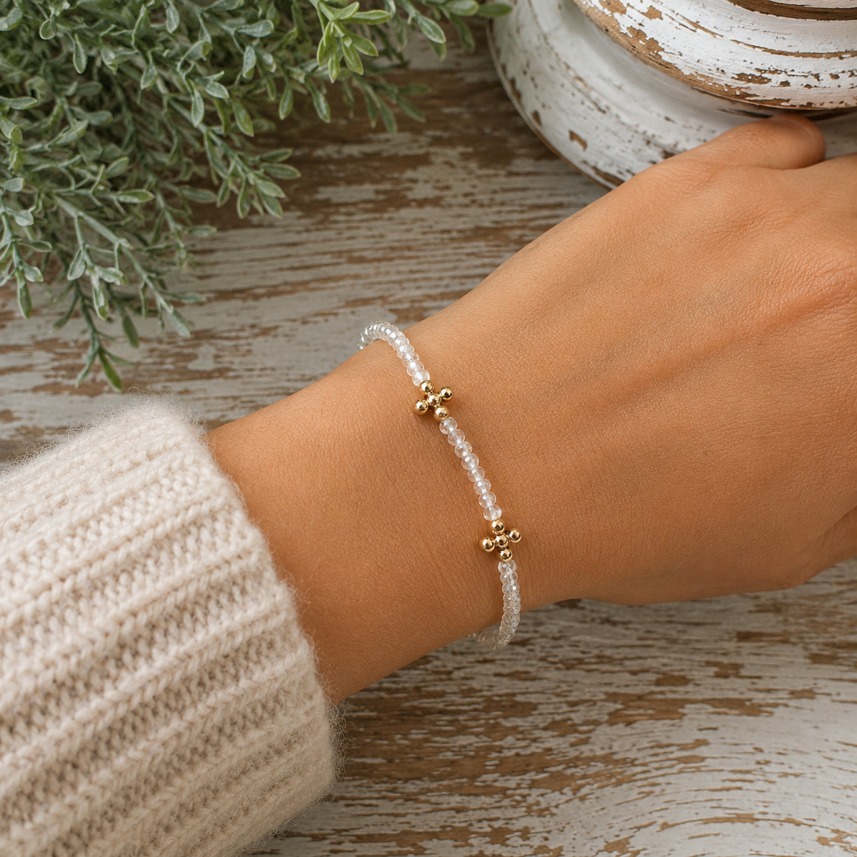 Woman wearing clear faceted beaded bracelet with gold cross accents on wrist with cream knit sweater and rustic chippy white pedestal backdrop
