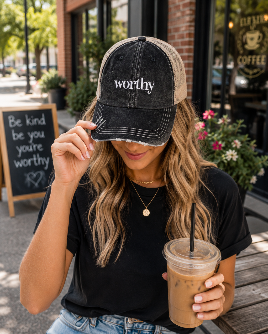 A woman wearing a distressed black and beige trucker hat with the word 'worthy' embroidered in white on the front, holding an iced coffee in front of a coffee shop storefront.