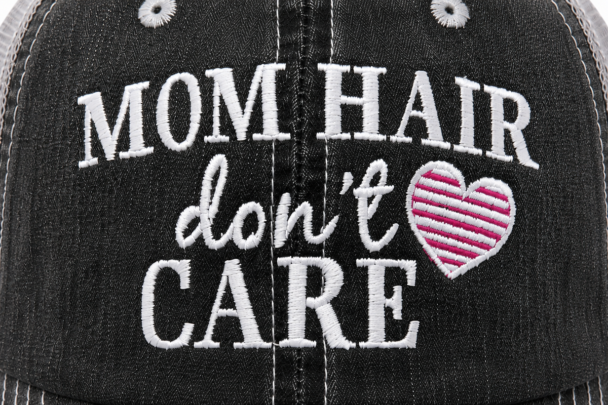 Close-up of Mom Hair Don't Care embroidery with pink striped heart detail on distressed charcoal trucker hat
