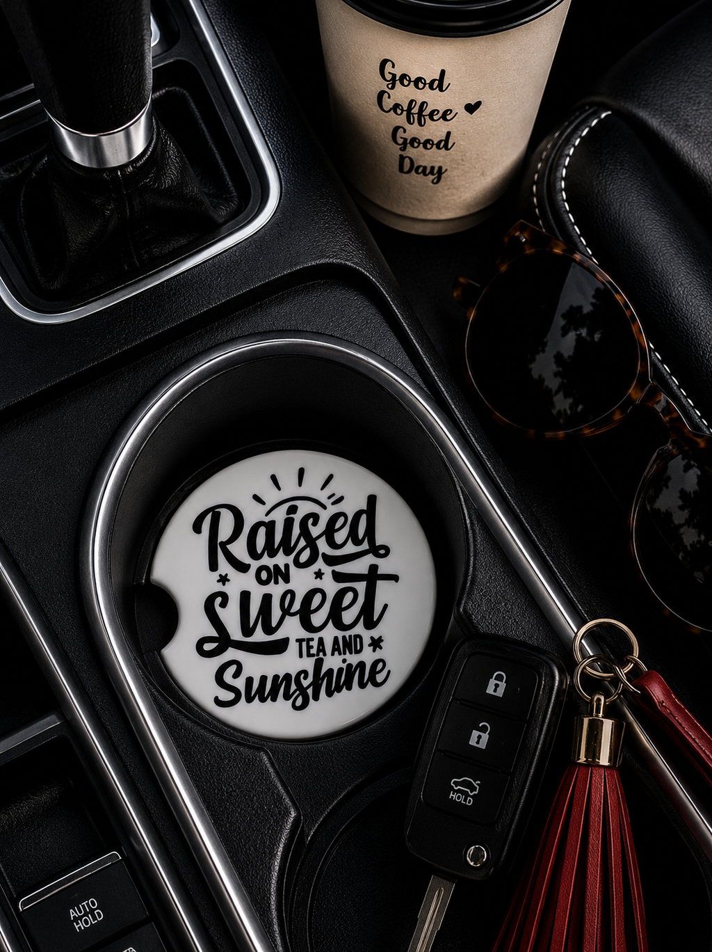 Raised on Sweet Tea and Sunshine ceramic car coaster in cup holder — bold black script on white, shown in car console with coffee cup, sunglasses, and red tassel keychain

