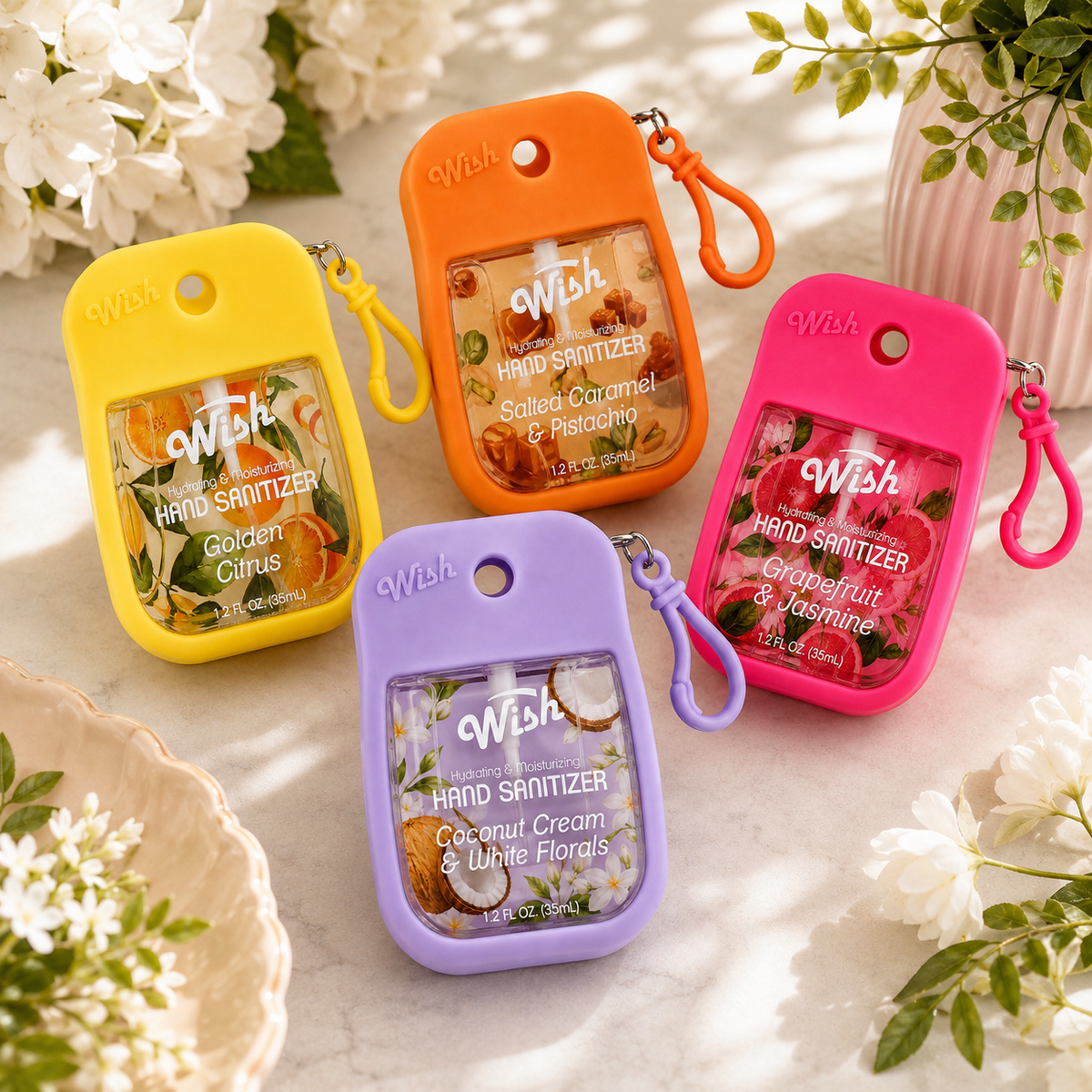 Four small Wish brand hand sanitizer keychain pouches in yellow, orange, pink, and purple are arranged on a light marble surface with white flowers and greenery around them. Each pouch has a different citrus and floral scent label visible through the clear front window.